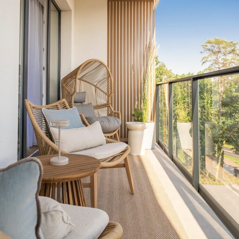 Prywatny balkon z rattanowymi fotelami, miękkimi poduszkami, małym stolikiem i szklaną balustradą oraz widokiem na drzewa i słońce.
