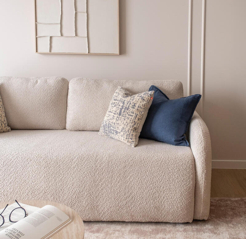 Kanapa bouclé w kremie z wzorzystą i granatową poduszką, abstrakcyjna grafika i stolik z otwartą książką i okularami — przytulny kącik