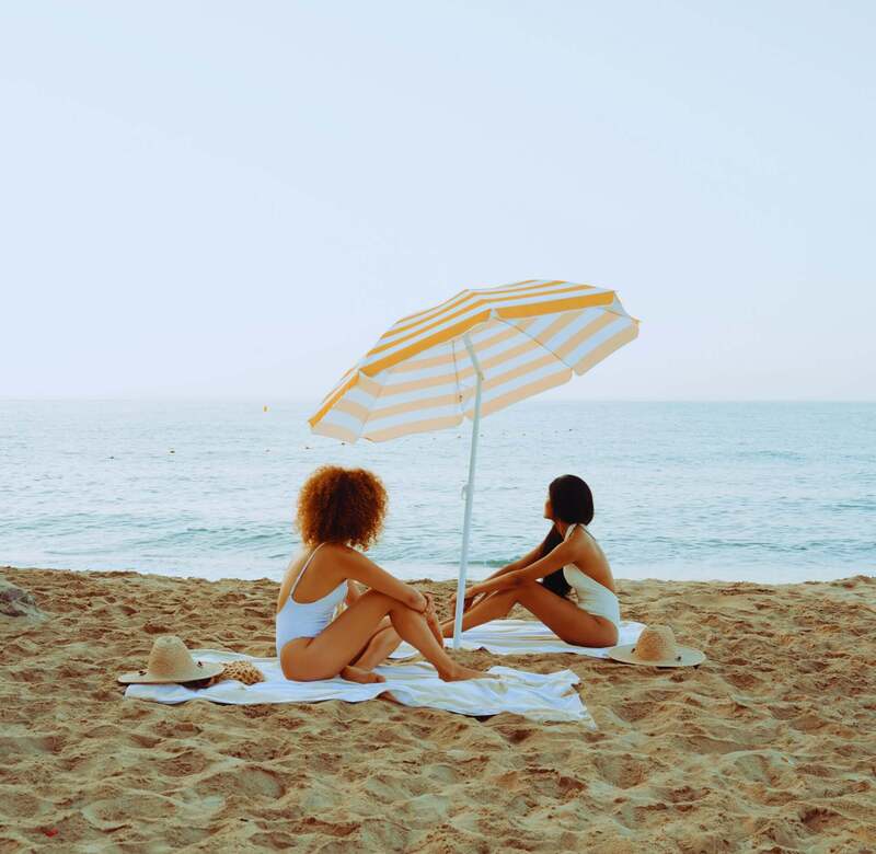 Dwie kobiety w strojach kąpielowych siedzą na ręcznikach pod żółto-białą parasolką na piaszczystej plaży, widok na spokojne morze.