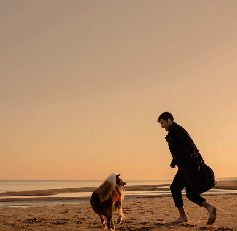 Gość boso bawiący się z psem na spokojnej plaży o zachodzie słońca, ciepłe złote światło, przyjazne zwierzętom nadmorskie otoczenie.