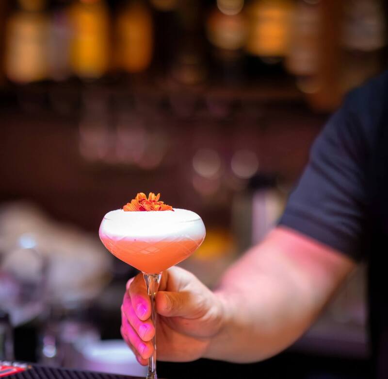 Koktajl w kieliszku coupe z pianką i jadalnym kwiatem trzymany przez barmana przy barze