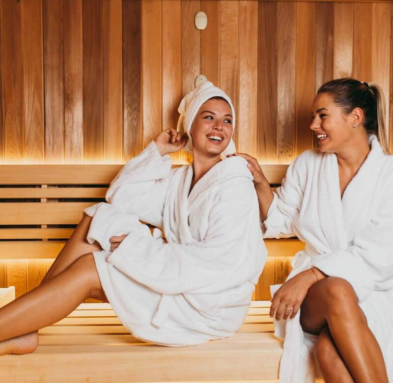 Dwie kobiety w białych szlafrokach relaksują się na drewnianych ławach sauny przy ciepłym, nastrojowym oświetleniu