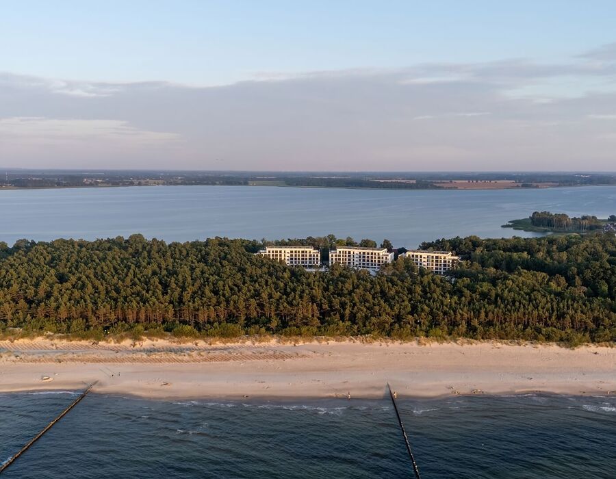 Nowoczesny nadmorski kompleks hotelowy schowany za sosnowym lasem, szeroka piaszczysta plaża z falochronami i spokojne morze o zmierzchu