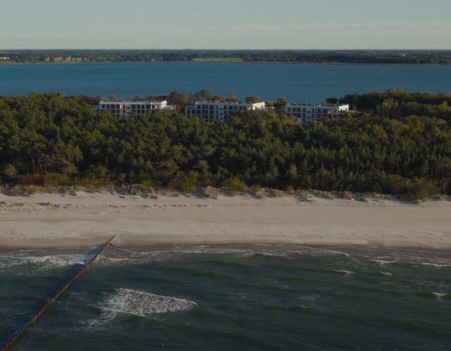 Nowoczesne niskie budynki hotelowe za sosnowym lasem, piaszczysta plaża z drewnianym falochronem i widokiem na morze, spokojny wypoczynek