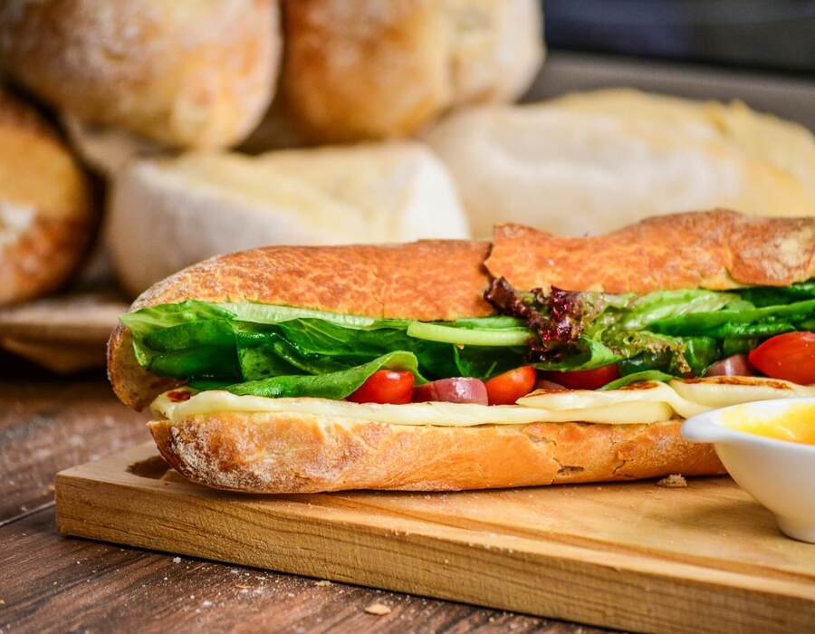 Świeża bagietka z roztopionym serem, chrupiącą sałatą i pomidorkami koktajlowymi na drewnianej desce, z sosem i bułkami w tle.