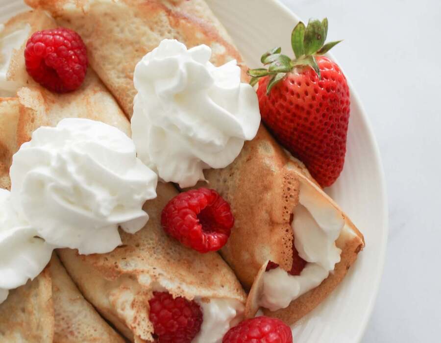 Delikatne naleśniki z waniliowym kremem i świeżymi malinami, udekorowane bitą śmietaną na białym talerzu