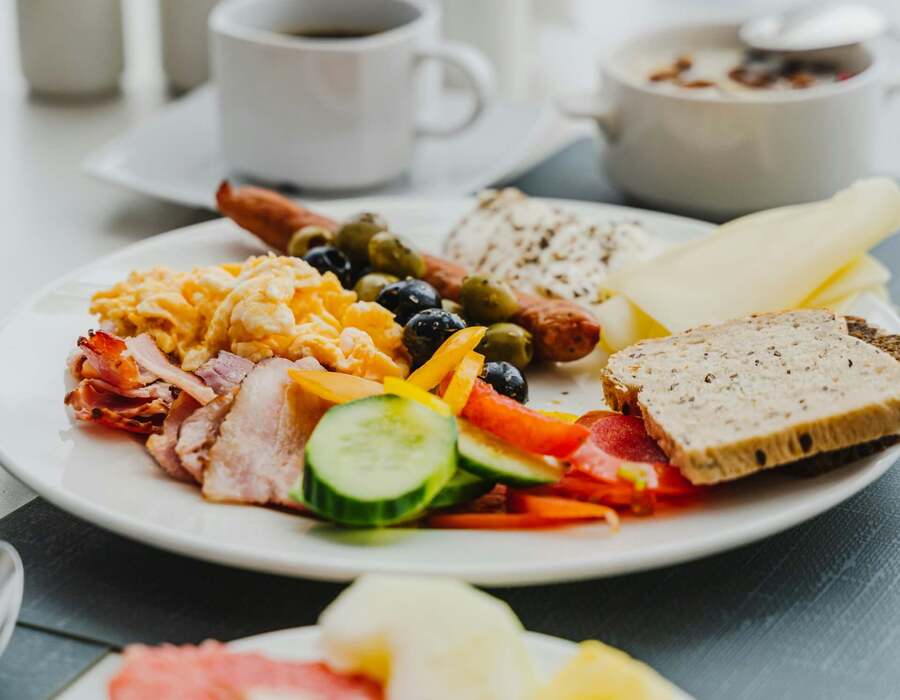 Śniadaniowy talerz z jajecznicą, boczkiem, kiełbasą, oliwkami, ogórkiem, serem, pełnoziarnistym chlebem, kawą i świeżymi owocami.