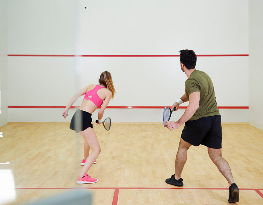 Dwójka gości gra w squasha na jasnym hotelowym korcie z drewnianą podłogą, przeszkloną ścianą i czerwonymi liniami