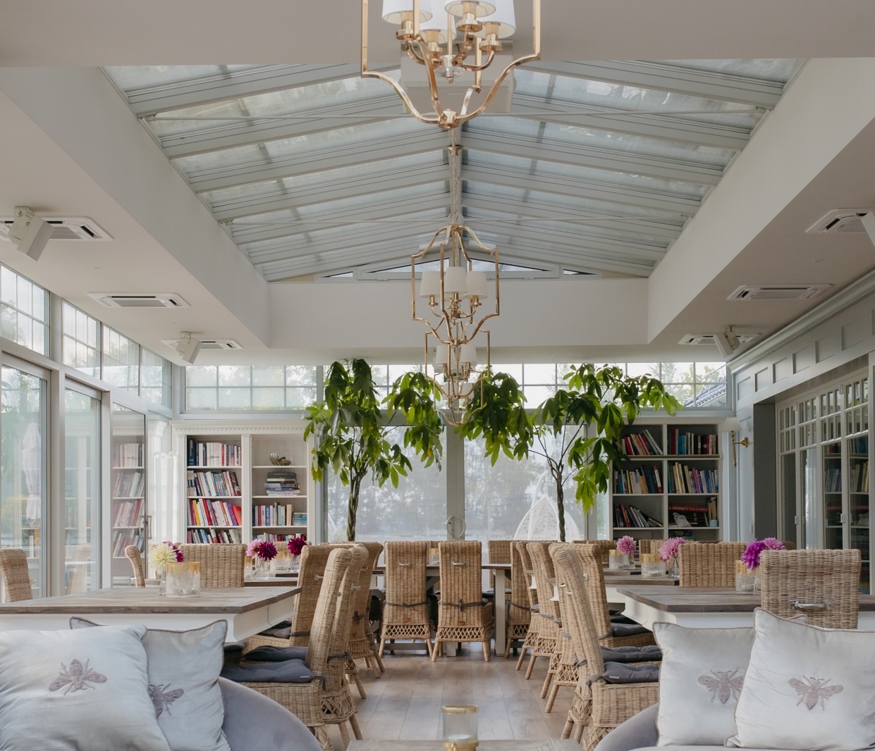 Jasna oranżeria z jadalnią i salonem — szklany świetlik, złote żyrandole, rattanowe krzesła, miękkie sofy, zielone pufy i regały z książkami.