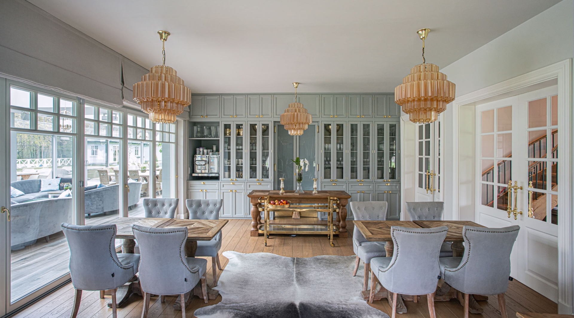 Elegancka jadalnia z pikowanymi szarymi krzesłami, drewnianymi stołami, przeszklonymi szafkami, mosiężnymi żyrandolami i dywanem ze skóry