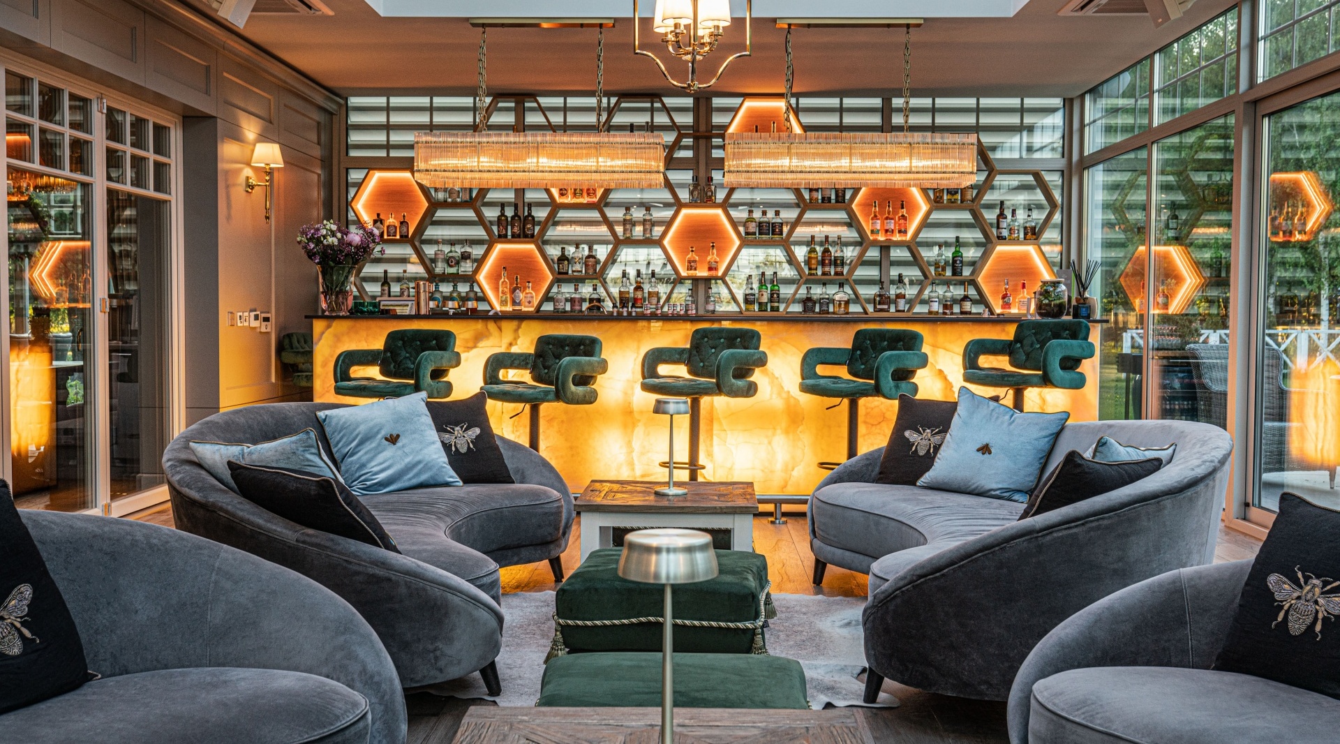 Hotelowy lounge z podświetlanymi sześciokątnymi półkami, barem z onyksu, zielonymi hokerami i szarymi sofami przy dużych oknach