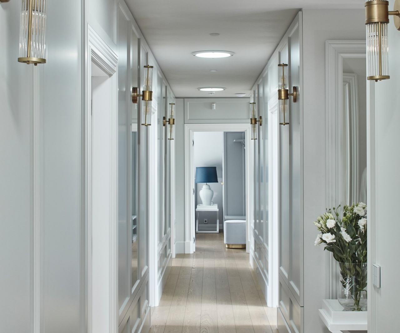Elegancki korytarz hotelowy z boazerią, mosiężnymi kinkietami, okrągłymi lampami, jasną drewnianą podłogą i stołem z kwiatami