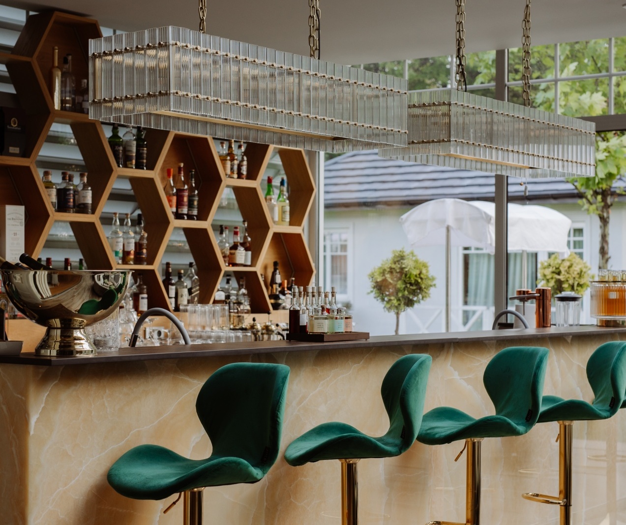 Luksusowy bar z marmurowym blatem, zielonymi hokerami na złotych podstawach, heksagonalnymi półkami z butelkami i żyrandolami.
