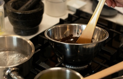 Ręka mieszająca roztopioną czekoladę w stalowej misce na kuchence gazowej; obok garnuszki z wrzącą wodą, masłem i drewnianymi łopatkami