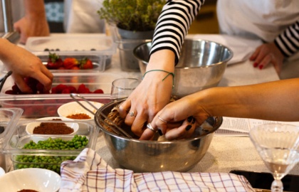 Goście w fartuchach na warsztatach kulinarnych mieszają ciasto w misce, obok pojemniki z groszkiem, truskawkami i ziołami.