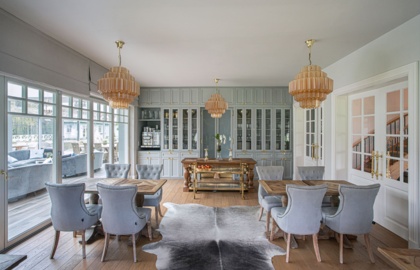 Elegancka jadalnia z pikowanymi szarymi krzesłami, drewnianymi stołami, przeszklonymi szafkami, mosiężnymi żyrandolami i dywanem ze skóry