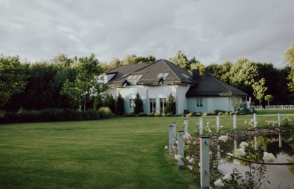 Biała wiejska willa z ciemnym dachem i oknami dachowymi, szeroki zadbany trawnik i staw obsadzony różami przy sznurze