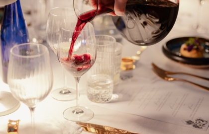 Ręka nalewa czerwone wino do kieliszka przy elegancko nakrytym białym stole ze złotymi sztućcami i małą przystawką