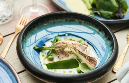 Grillowana dymka i małż św. Jakuba z groszkiem w kremowym sosie w niebieskiej ceramicznej misce, złote sztućce i kieliszek czerwonego wina