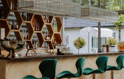 Bar hotelowy z zielonymi aksamitnymi hokerami, marmurowym blatem, sześciokątnymi półkami na alkohole i szklanymi lampami, widok na ogród.