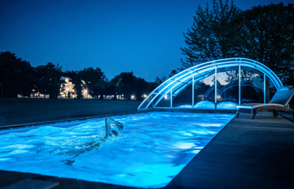 Nocny basen na zewnątrz z niebieskim oświetleniem, gość płynie kraulem przy zakrzywionej, przesuwnej szklanej kopule, leżak i oświetlony hotel w tle.