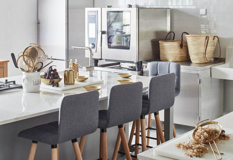 Otwarta kuchnia hotelowa z wielofunkcyjnym piecem, białymi płytkami, kwarcową wyspą i trzema szarymi hokerami.