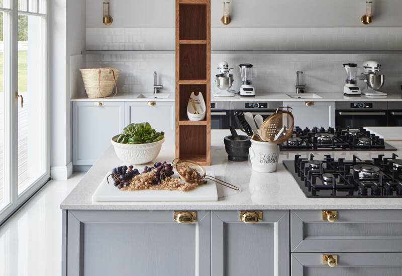 Jasna kuchnia z dużą szarą wyspą, płytą gazową, mosiężnymi uchwytami, drewnianą półką, robotami kuchennymi i misą z owocami i sałatą.