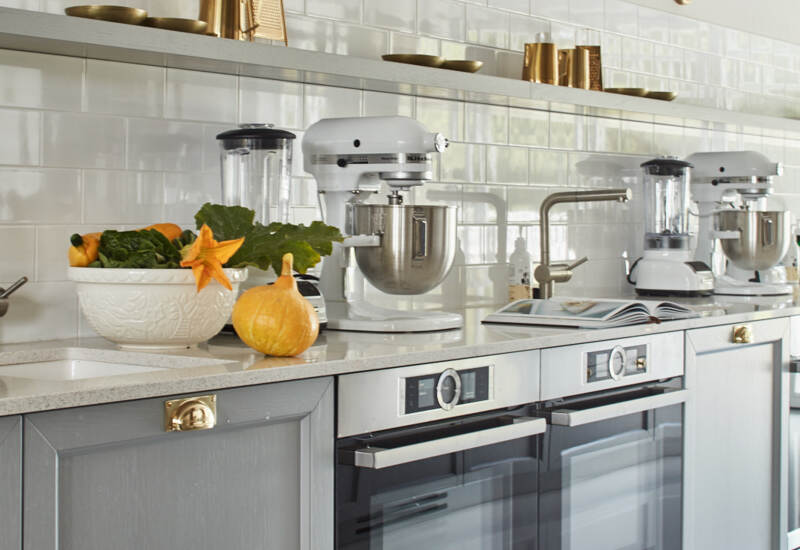Kuchnia z szarymi szafkami, podwójnymi piekarnikami, mikserami i blenderem, białymi płytkami typu metro i złotymi akcentami.