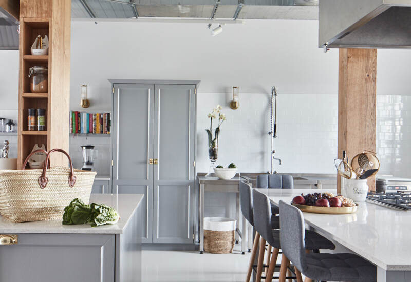 Jasna nowoczesna kuchnia z szarą wyspą i marmurowym blatem, czterema szarymi hokerami, odsłoniętym kanałem i drewnianymi belkami.