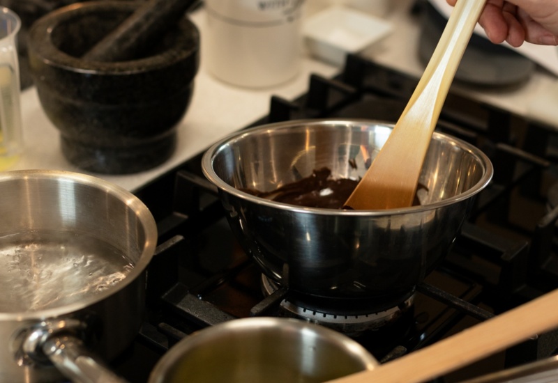 Ręka mieszająca roztopioną czekoladę w stalowej misce na kuchence gazowej; obok garnuszki z wrzącą wodą, masłem i drewnianymi łopatkami