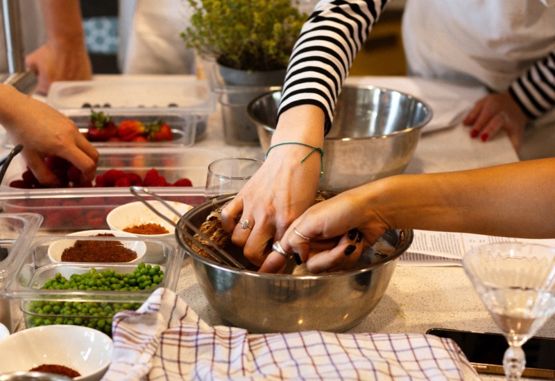 Goście w fartuchach na warsztatach kulinarnych mieszają ciasto w misce, obok pojemniki z groszkiem, truskawkami i ziołami.