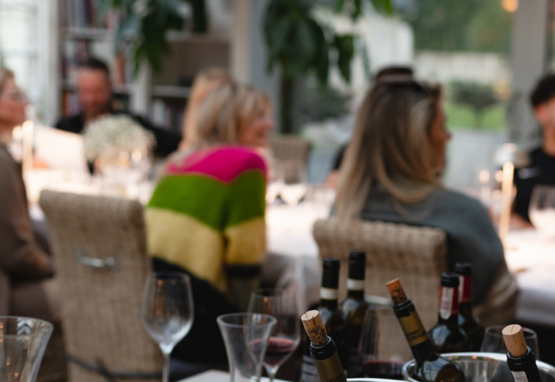 Butelki wina w metalowych wiaderkach i dekanter na stoliku degustacyjnym, goście przy świecach w przeszklonym salonie z wiklinowymi krzesłami.