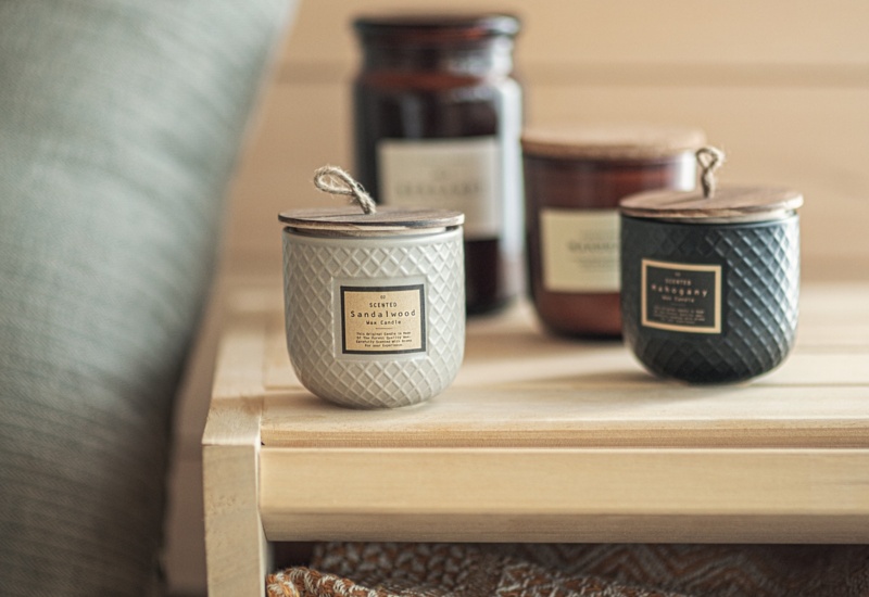 Drewniana półka z zapachowymi świecami w szklanych słoikach, złożonym wzorzystym kocem i miękką poduszką tworząca przytulny kącik w klimacie spa.