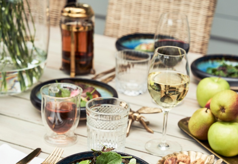 Stół z przystawką z łososia w niebieskiej misce z zieleniną, kieliszek białego wina, zaparzacz tłokowy, wyplatane krzesła i miska jabłek.