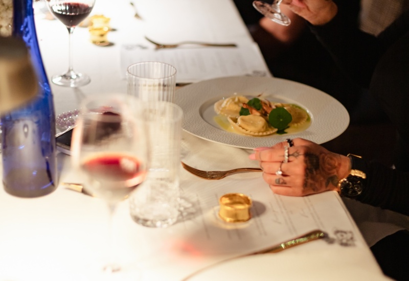 Długi stół z białym obrusem, ravioli na talerzach, goście trzymają kieliszki czerwonego wina, ciepła nastrojowa atmosfera