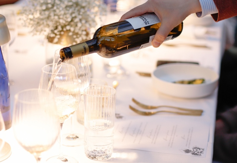 Ręka wlewa białe wino do kieliszka przy świecach, biały obrus, bukiet gipsówki, przystawka ze szparagów i złote sztućce