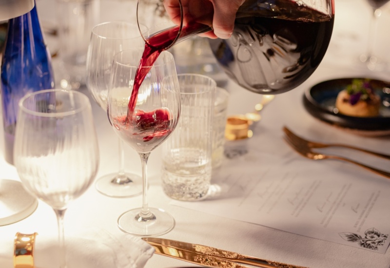 Ręka nalewa czerwone wino do kieliszka przy elegancko nakrytym białym stole ze złotymi sztućcami i małą przystawką