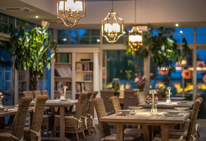Przytulna restauracja hotelowa z drewnianymi stołami nakrytymi do kolacji, rattanowymi krzesłami, ciepłym oświetleniem i wazonikami przy oknach.