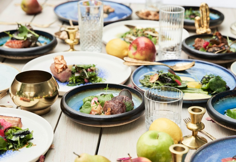 Wspólne degustacyjne dania na niebieskiej ceramice, świeże owoce, kryształowe szklanki i mosiężne świeczniki na stole