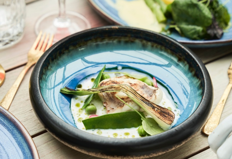 Grillowana dymka i małż św. Jakuba z groszkiem w kremowym sosie w niebieskiej ceramicznej misce, złote sztućce i kieliszek czerwonego wina