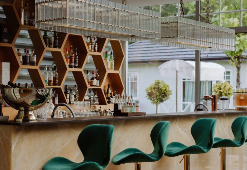 Bar hotelowy z zielonymi aksamitnymi hokerami, marmurowym blatem, sześciokątnymi półkami na alkohole i szklanymi lampami, widok na ogród.