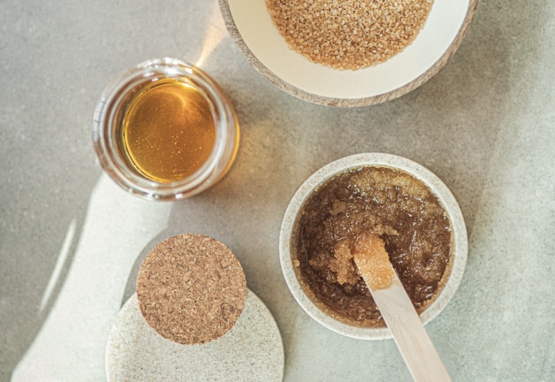 Miska z grubym brązowym cukrem, słoik złotego oleju i miseczka z peelingiem oraz drewniana szpatułka — naturalny zestaw spa.