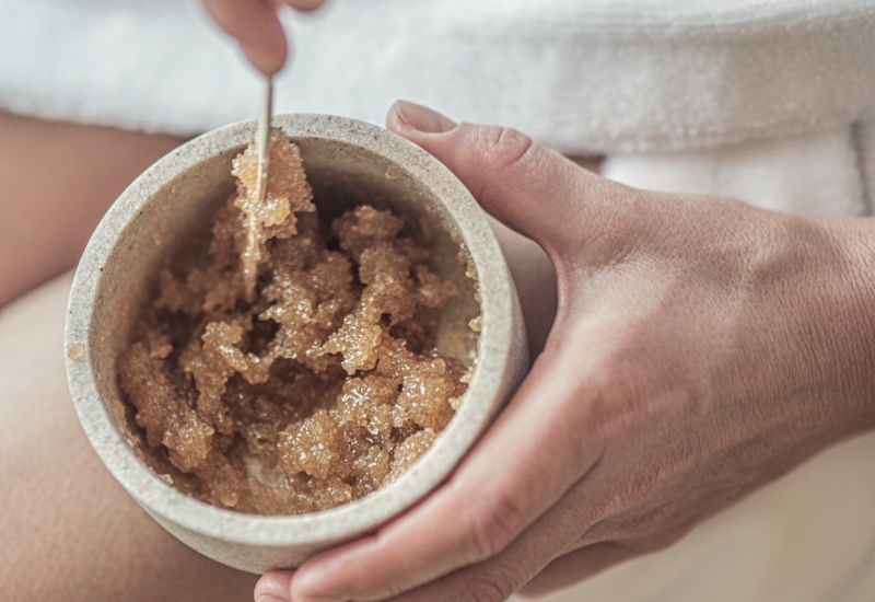 Ręce nabierające brązowego peelingu cukrowego z kamiennej miseczki, gość w białym szlafroku przed zabiegiem spa