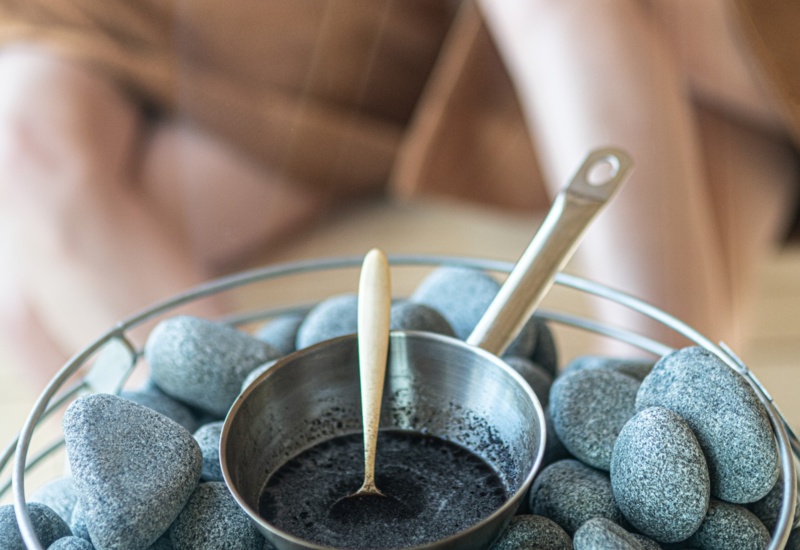 Metalowa miseczka z ciemnym peelingiem i łyżką na gładkich kamieniach sauny, w tle rozmyte osoby w szlafrokach