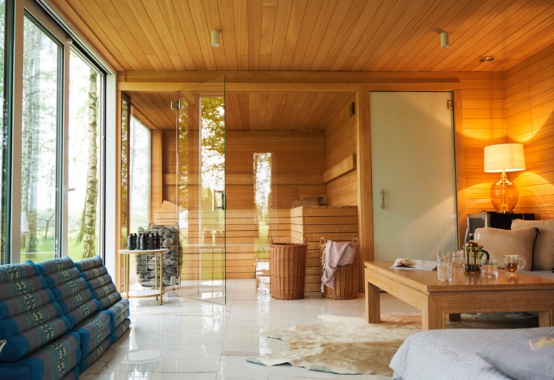 Sauna z drewnianym wnętrzem i przeszklonymi drzwiami obok przytulnego salonu, widok na las, wiklinowe kosze i herbata.