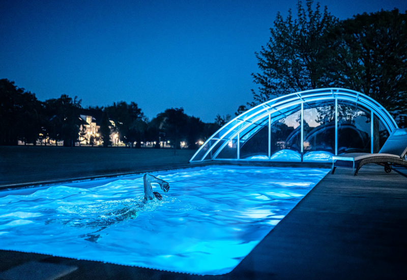 Nocny basen na zewnątrz z niebieskim oświetleniem, gość płynie kraulem przy zakrzywionej, przesuwnej szklanej kopule, leżak i oświetlony hotel w tle.