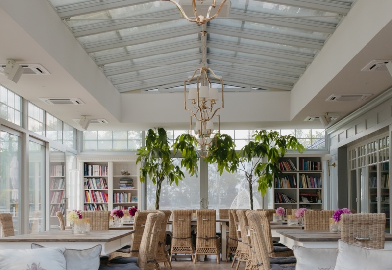 Jasna oranżeria z przeszklonym dachem, złotymi żyrandolami, wiklinowymi krzesłami, zielonymi drzewkami, miękkimi sofami i regałami.