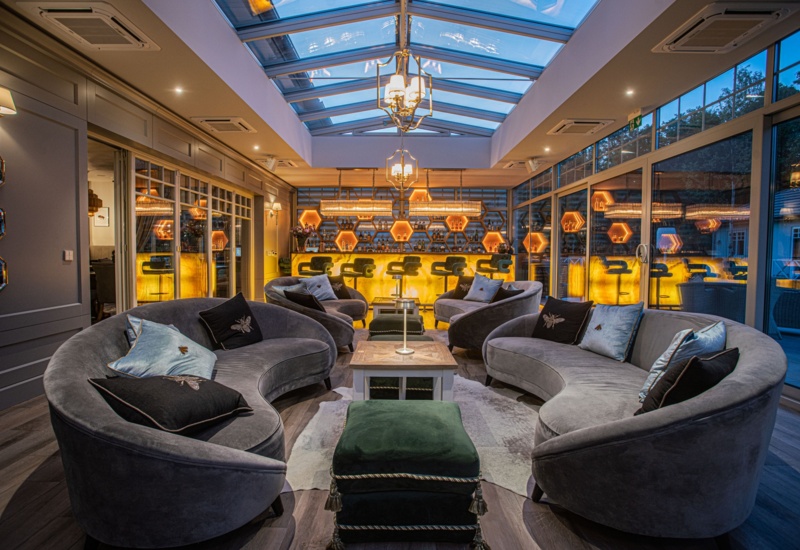 Luksusowy salon hotelowy z zaokrąglonymi aksamitnymi sofami, przeszklonym świetlikiem i podświetlanym sześciokątnym barem.