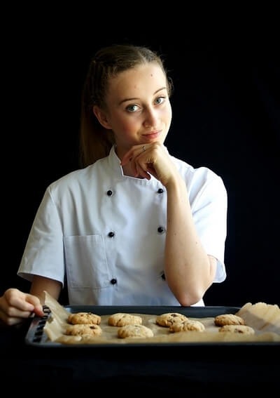 Hotelowa cukierniczka w białym fartuchu trzyma blachę świeżo upieczonych ciastek, prezentując ofertę cukierni i deserów.