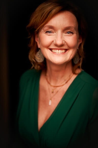 Uśmiechnięta kobieta w eleganckiej zielonej sukni, nosi kilka naszyjników i ozdobny kolczyk, ciepły portret